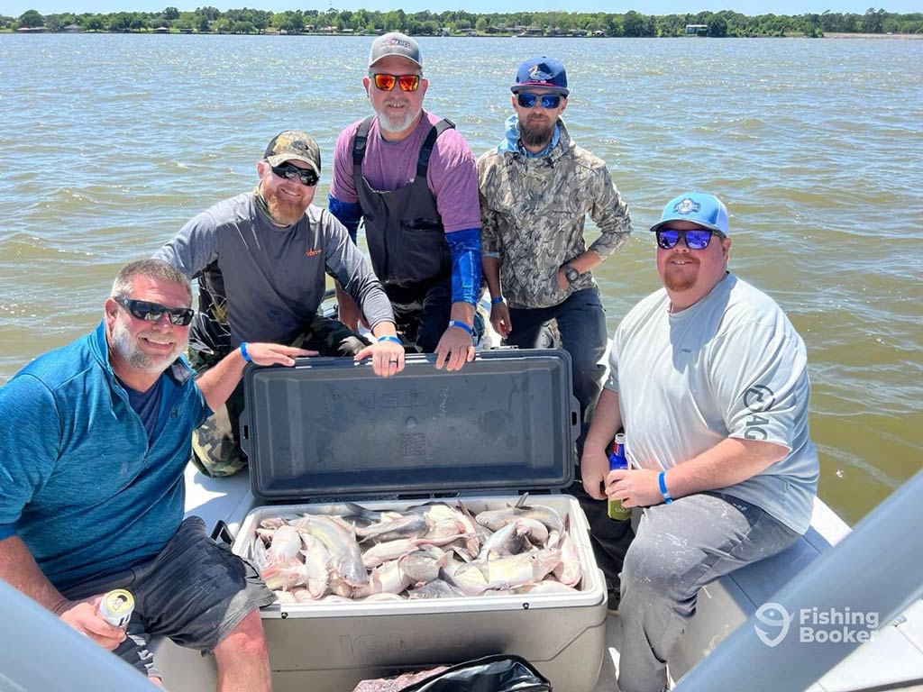 A group of happy anglers aboard a fishing charter in Lake Livingston on a sunny day, sitting around a cooler full of fish, with the water visible behind them and land in the distance