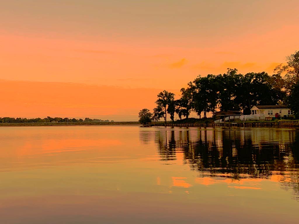 A view across the calm waters of Lake Livingston at sunset, with the sun creating a bright orange hue on the horizon, with a tree-lined shore visible as a silhouette in the distance