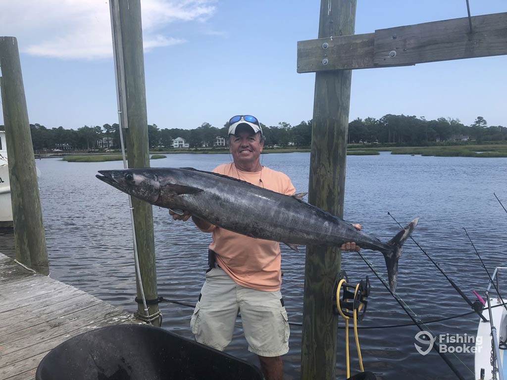 A man on a dock after a trip offshore from Calabash, holding a large, dark Kingfish with both hands with inshore waters visible behind him