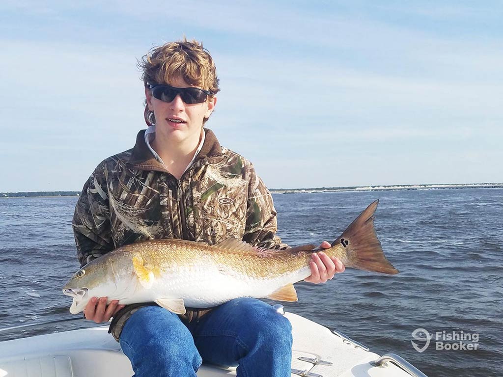 A young man in a khaki jacket and sunglasses, sitting on a fishing charter in Calabash, NC, with a Redfish on his lap with the water visible behind him