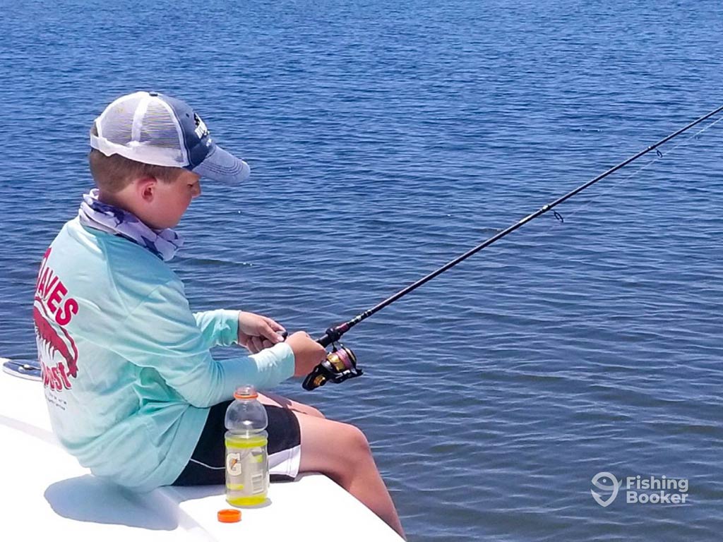 A young boy sitting over the side of an inshore fishing charter in North Carolina, casting his fishing rod into the water on a clear day