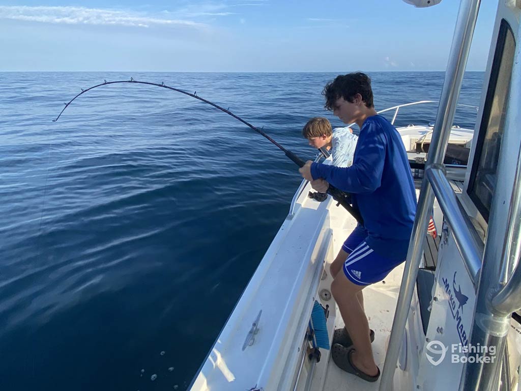 A teenage boy fishing over the side of a charter boat in deep waters, with his rod bent on a sunny day