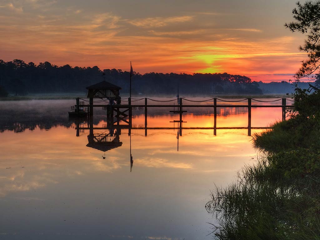 A view across the calm waters of the Calabash River at sunset, with a small wooden fishing deck in the foreground and the sun visible behind some trees in the distance