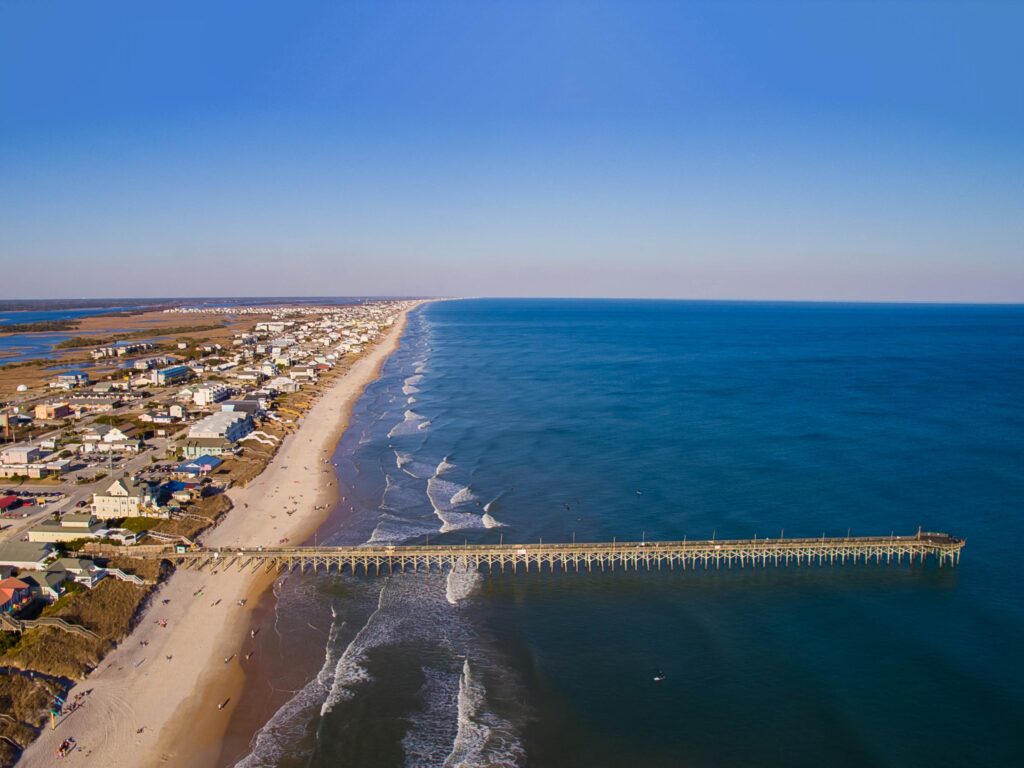 An aerial view of a town near the Little River Inlet on a clear day with a fishing charter sticking out into the ocean on the right of the image and the Intracoastal Waterway visible on the left of the image