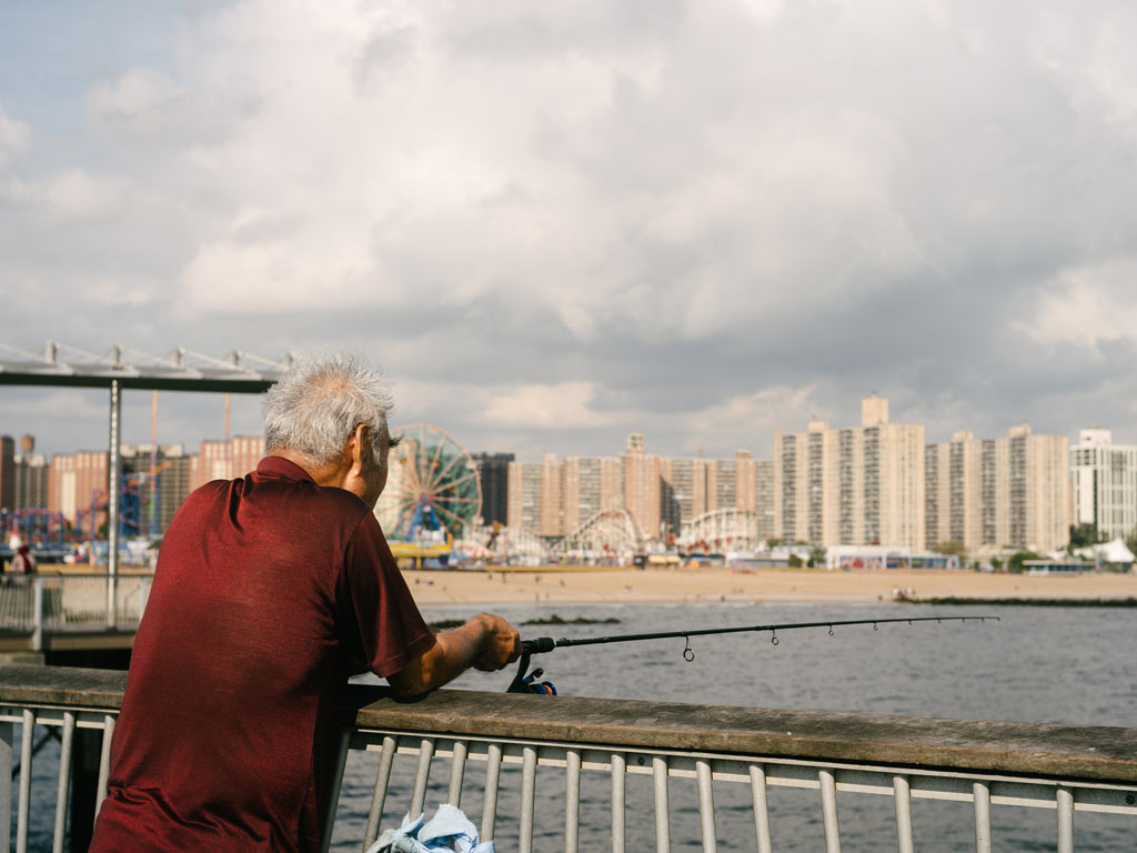 A elderly man in a red shirt fishing over a fence on Coney Island Pier in New York, with the water in front of him and buildings visible across on the shore.