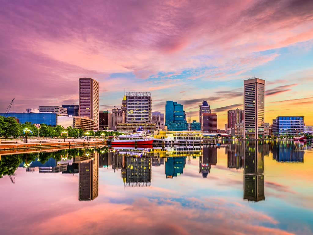 A view of the skyline of Baltimore at sunset with the city's buildings and the dramatic pinkish skies reflecting off the water surface in the foreground.