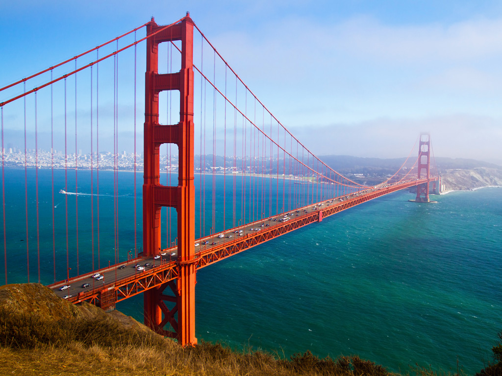 A view of the Golden Gate Bridge in San Francisco, California from a hill on a clear day, with striking blue waters flowing underneath the bridge and the city visible in the distance.