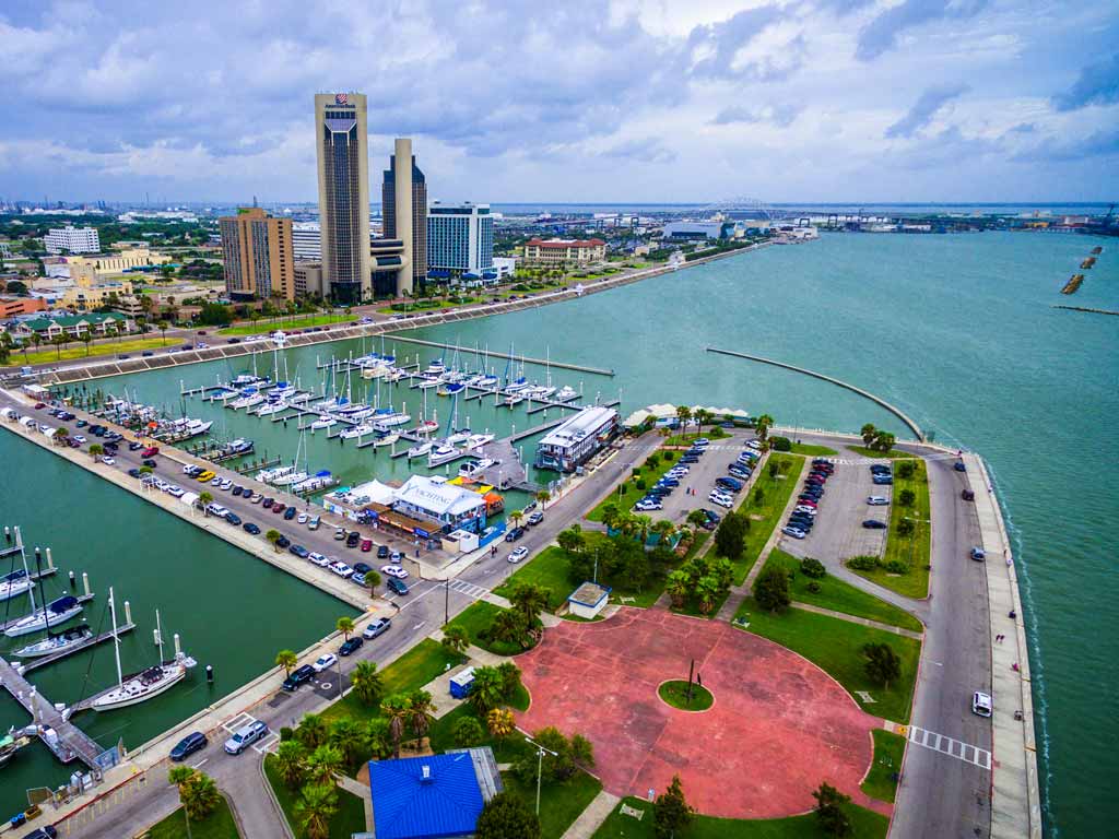 An aerial view of the city of Corpus Christi, home to some of the best fishing in Texas, and the Corpus Christi Marina with various docked boats and the Norma Urban Park visible on a cloudy day.