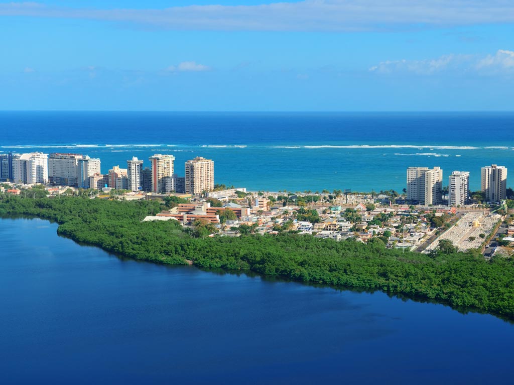 An aerial view of San Juan and its buildings, taken somewhere close to the Martin Peña channel, with the inshore waters visible in the foreground and the open ocean and clear skies stretching out in the background beyond the city.