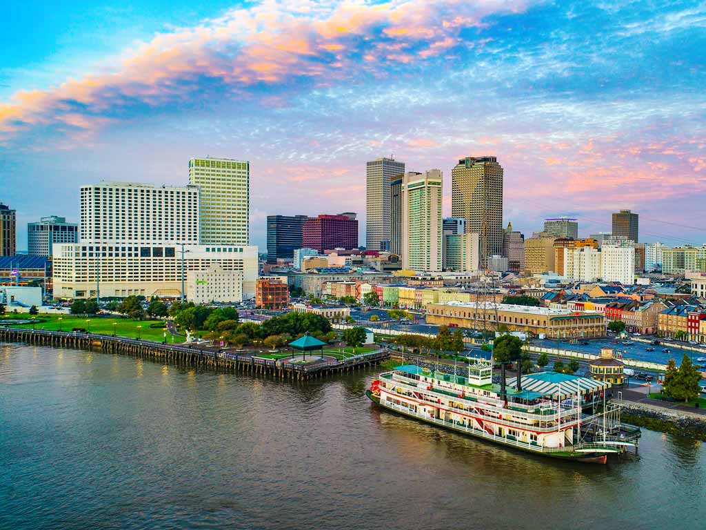 A sunset view of the New Orleans skyline taken from above the water, with the city's buildings visible against vividly-colored skies, and with a large ship in the water.