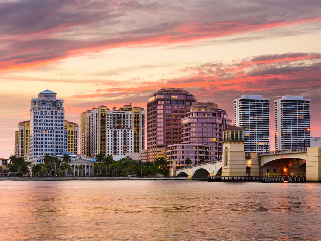 A sunset shot of the skyline of West Palm Beach, with the setting sun giving the entire photo an orange-pink hue, with waters visible in the forefront and buildings in the distance.