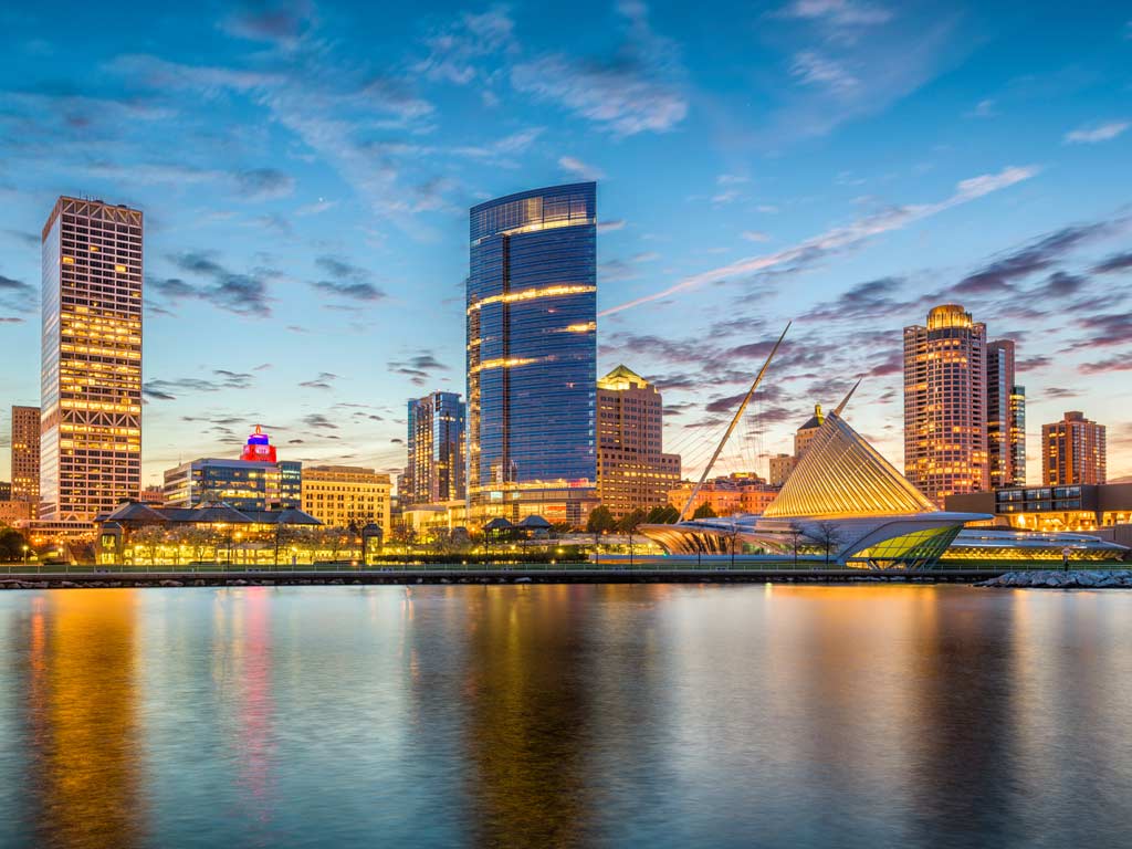 An evening shot of the waterfront of Milwaukee, taken from Lake Michigan,  with calm waters and lit-up buildings visible and partially-reflected in the water. 