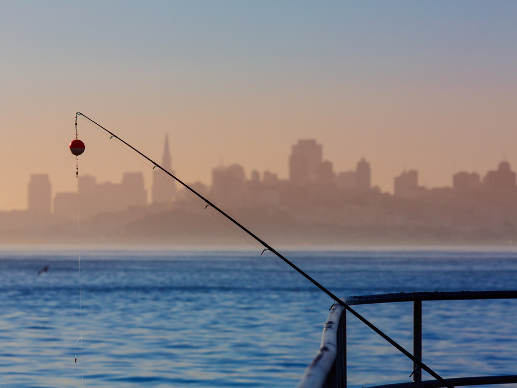 A fishing rod set against a fence, with a bobber and the line hanging down into the water with the foggy skyline of San Francisco in the background.