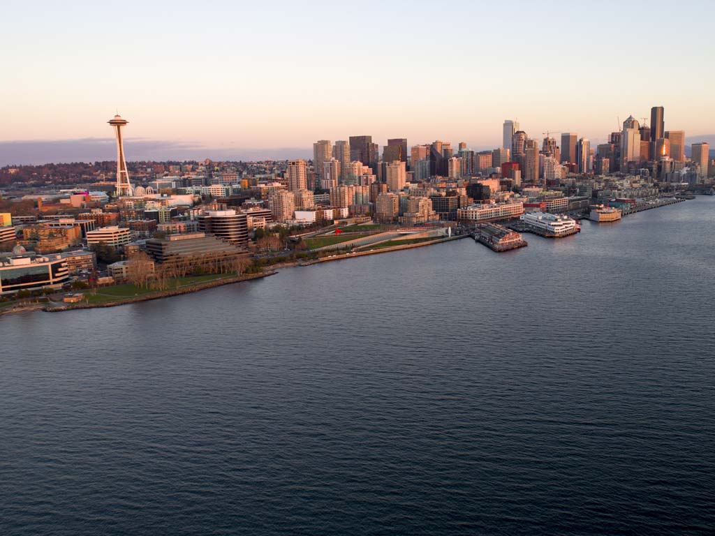 An aerial view of Seattle taken from above the Puget Sound, with the city's most famous landmark, the Space Needle, visible along with numerous other buildings at sunset on a clear day.