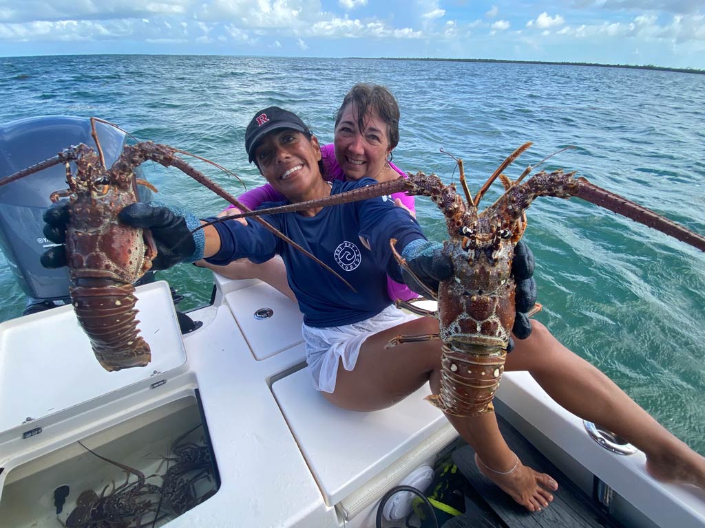 A photo of two happy anglers sitting on a charter boat while one of them is happily posing for the camera with a Lobster in each hand