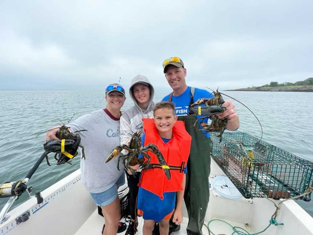 A group of anglers including a captain, a mom, and two sons standing on a charter boat and posing with several Lobsters they’ve caught during their season but on a cloudy and rainy summer day