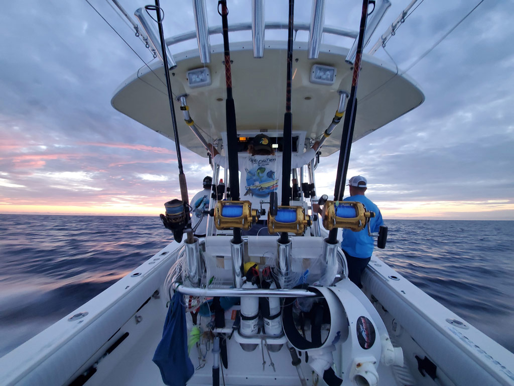 A photo of a rack of fishing rods, reels, a fighting belt, and additional gear and equipment, along with a captain with a first mate navigating their charter fishing boat before sunrise 