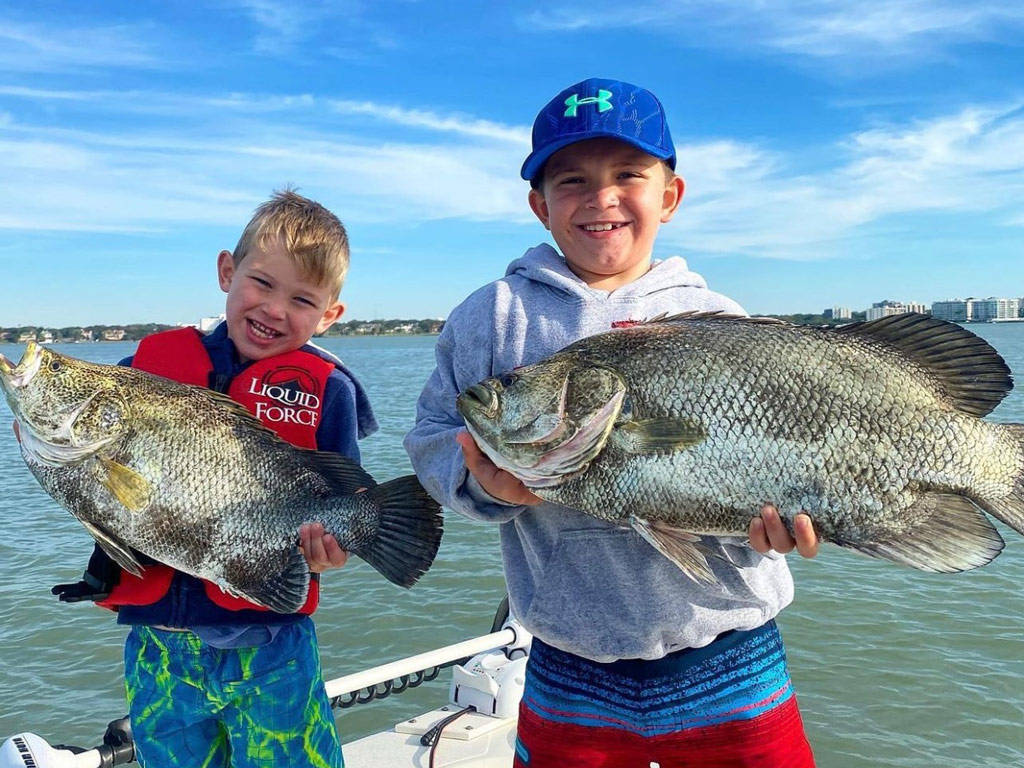 A photo of two happy kids standing on a charter boat in the Gulf near Florida’s Sports Coast and posing with two Tripletail fish they caught on their winter fishing trip