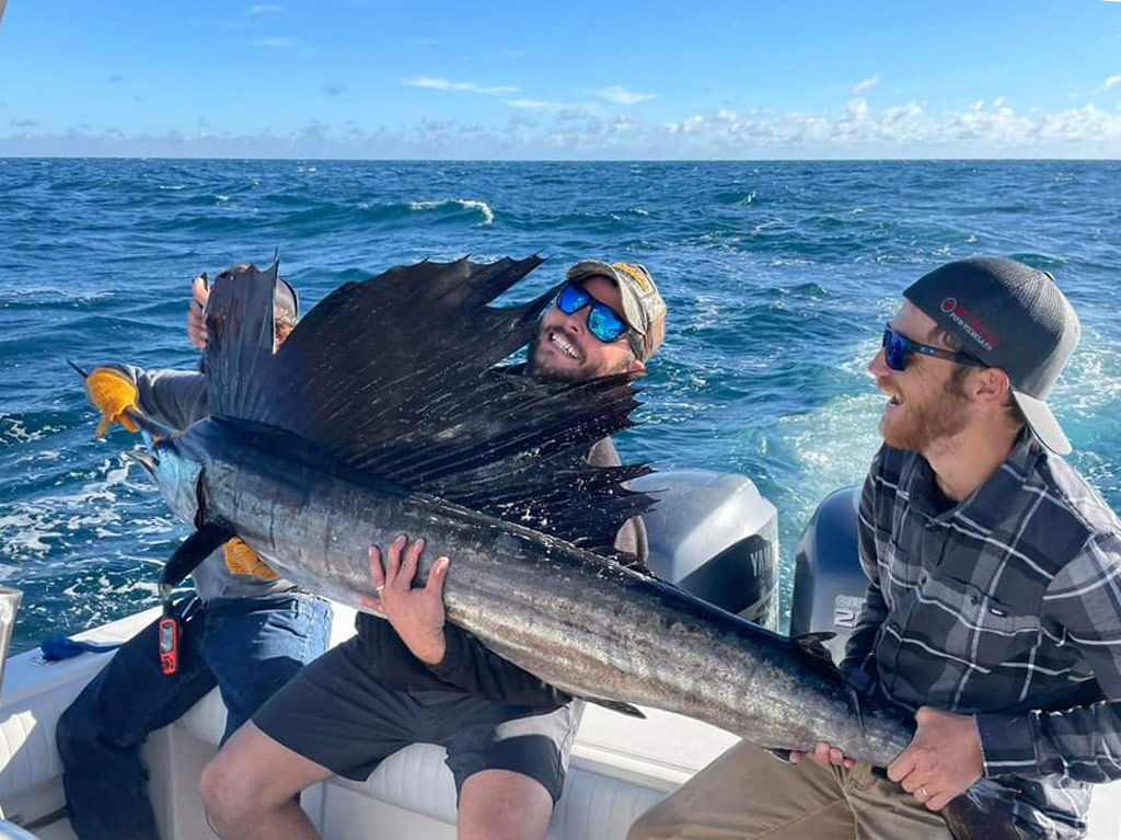 A photo of three happy anglers sitting on a charter boat and smiling while trying to hold a big Sailfish caught while deep sea angling out of Daytona Beach