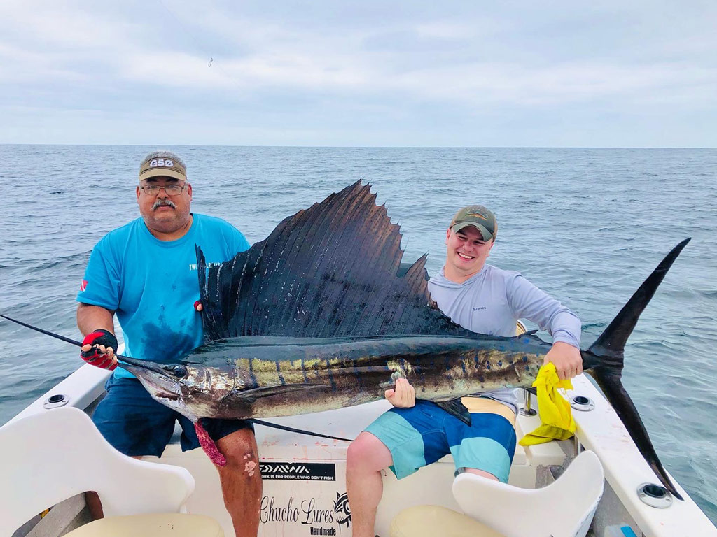 A father and a son sitting on a Puerto Vallarta charter fishing boat and struggling to hold a huge Sailfish with both hands while posing for a photo during their winter angling trip