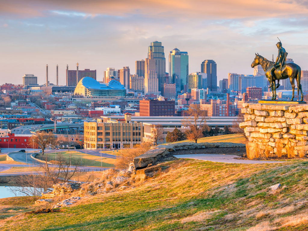 A sunrise view of Kansas City and its iconic skyline with skyscrapers, business buildings, park, pond, and the famous statue overlooking the city 