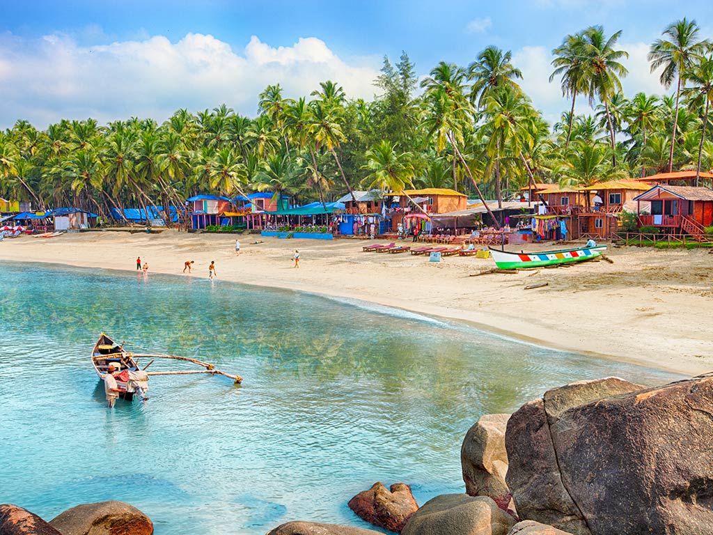 A view of a beach in a fishing village in India with brightly-colored shacks in front of palm trees on the shroe and a couple of boats visible in the water on a clear day