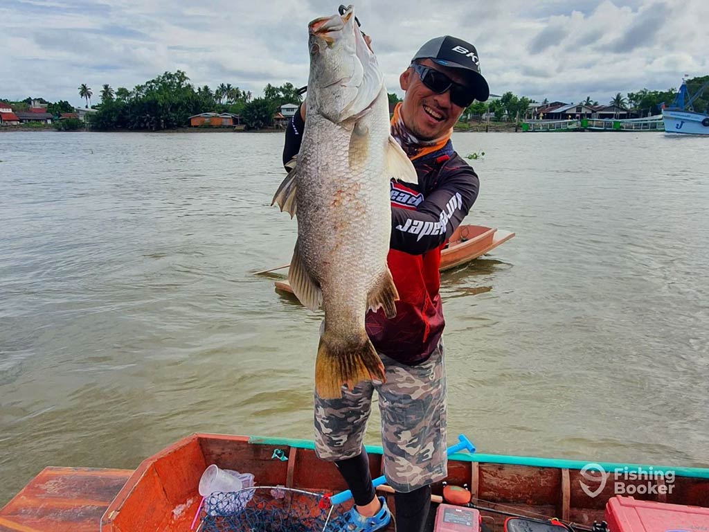 A man standing on a fishing boat on an inshore river in Asia holding a Barramundi upright with murky waters visible behind him, along with a tree-lined shore in the distance on a cloudy day