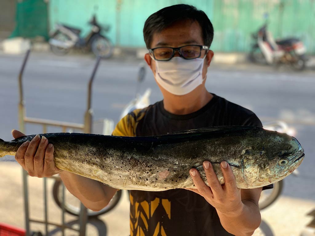 A man in glasses and a face mask holding an Indian Salmon while back on shore after a successful fishing trip, with the background blurred behind him