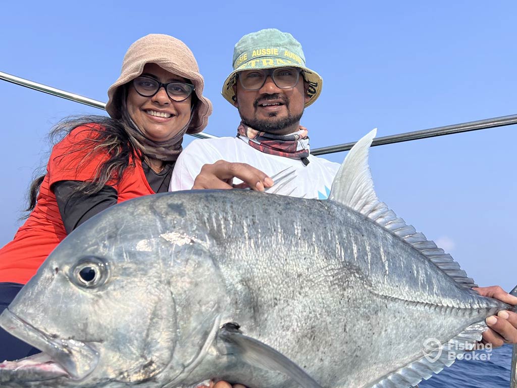 A man and a woman wearing bucket hats on a fishing boat in India with a large Giant Trevally across their laps on a clear day