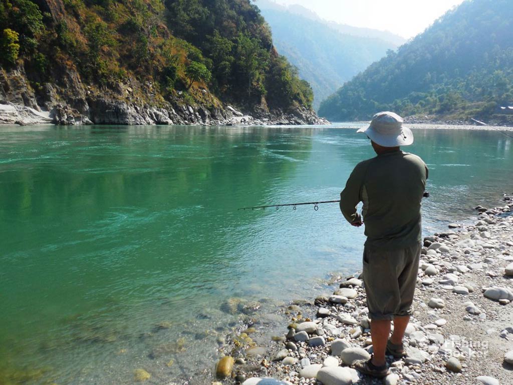 A view from behind of a man fishing into a freshwater lake high up in the mountains of India on a clear day, with crystal clear waters and rocky hills visible all around him