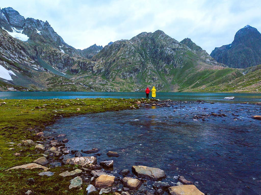 A view from a distance of two anglers in rain coats fishing in a mountain-top lake in the Himalayas in India, with snow visible on the peak of a mountain on the left of the image and dark blue water visible among the rocks in the foreground