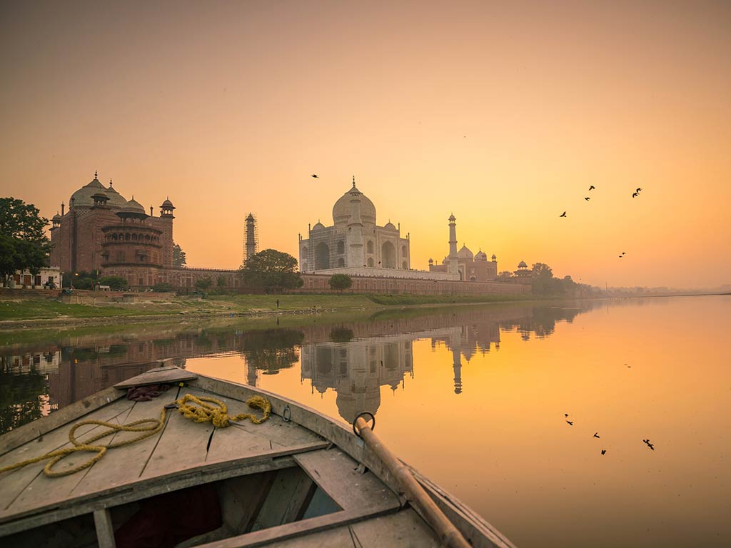 A view across the calm waters of a river from a boat towards the Taj Mahal at sunrise, with the sun visible behind the mausoleum in the distance and some birds flying on the right of the image