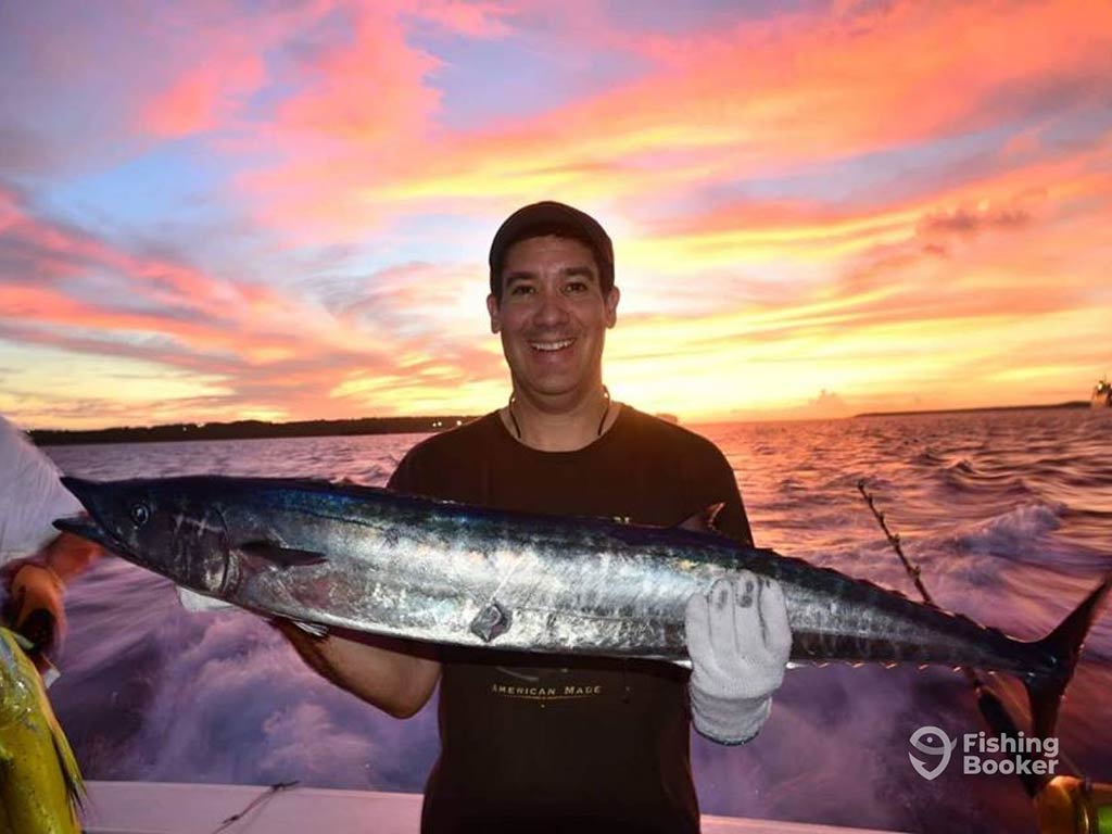 A man on a fishing charter in Guam holding a large Wahoo with the sun setting across the deep waters behind him