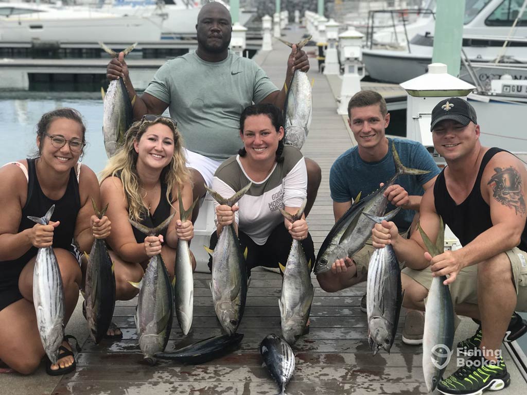 A group of three male and three female anglers crouching on a dock in Guam after a successful trip, with each holding a couple of Tuna