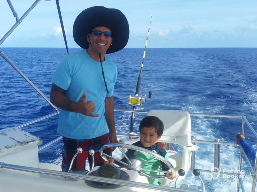 A man in a large hat stands next to a child who's mimicking controlling a deep sea fishing boat in Guam, with one trolling rod and the wake of the boat visible behind them