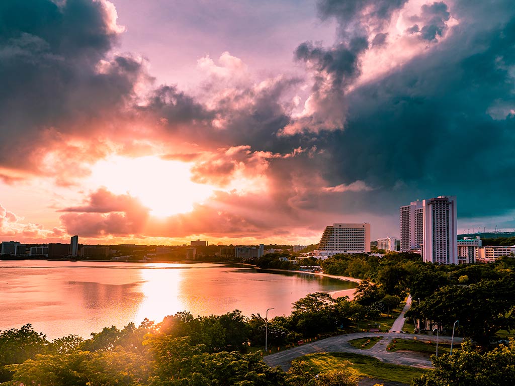 A view from a hill towards a beach and a bay in Guam at sunset, with the sun setting in the distance on the left of the image and high-rise buildings visible on the right