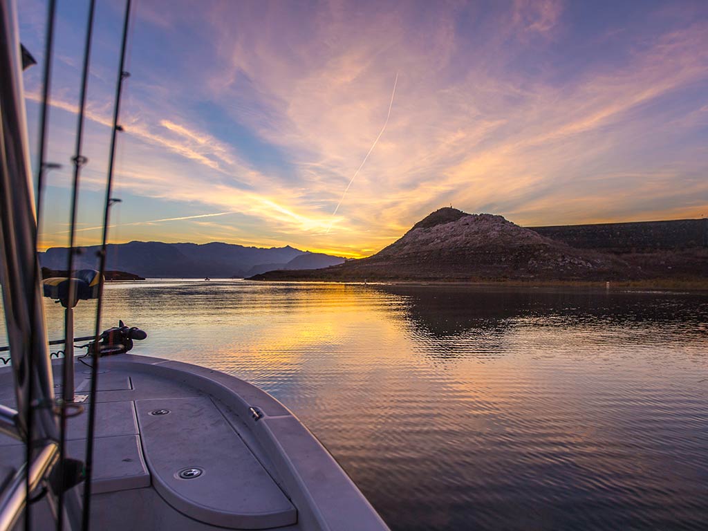 A view across the bow of a fishing boat on the calm waters of Lake Mead, Nevada, at sunset, with the sun visible setting behind some clouds in the distance