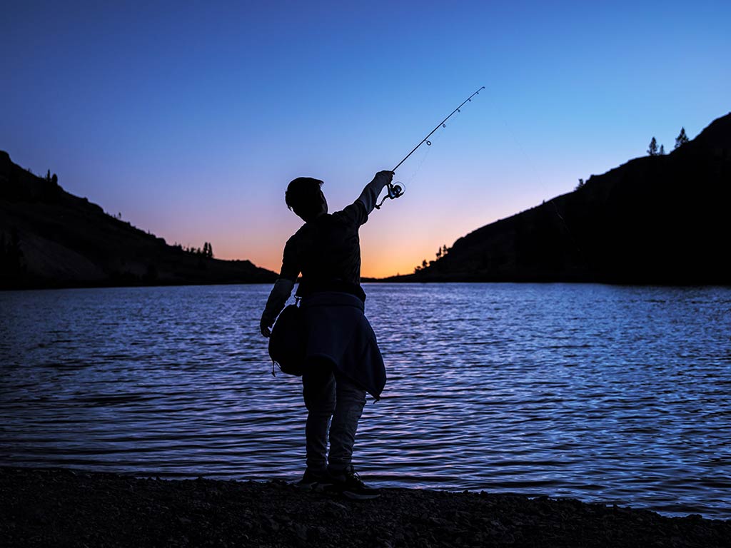 A view from behind of a silhouette of a boy fishing against the backdrop of a lake in Nevada at sunset with the sun having just disappeared in the distance