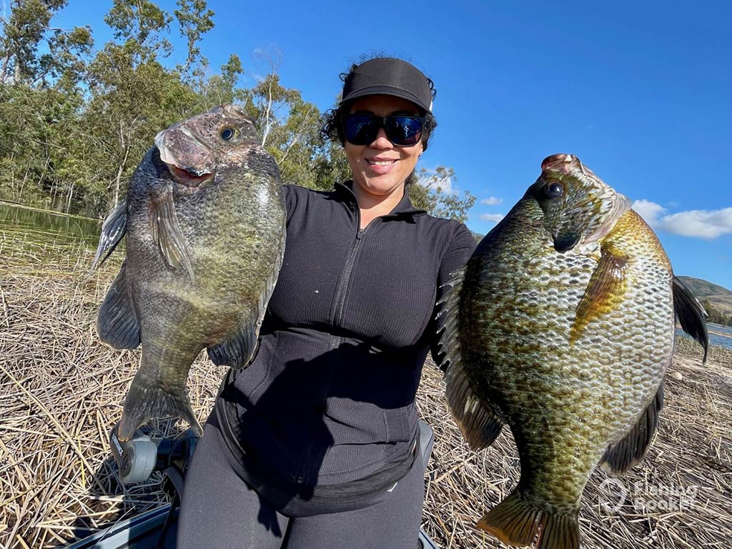 A closeup of a woman in sunglasses and a baseball cap holding two large Panfish on dry land next to a lake on a sunny day