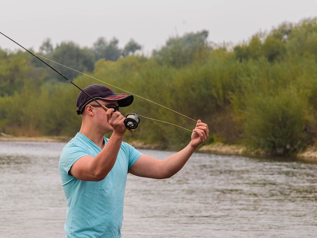 A man in mid-action, casting a fly fishing rod on a river in Nevada, with a green shoreline visible in the distance on a cloudy day