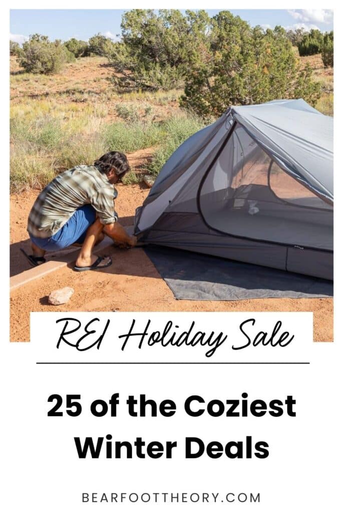 Pinterest image with text that reads "REI Holiday Sale 25 of the coziest winter deals." Image shows a man putting a tent stake into the ground to secure his gray tent.