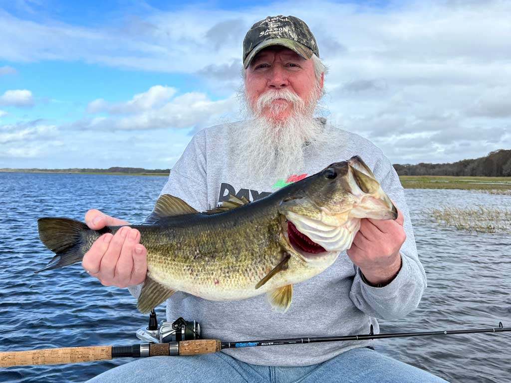 An angler in a hat and with a long white beard holding a sizeable Largemouth Bass he caught fishing on Lake Tohopekaliga in Florida, where it's common to use live bait to target these fish.