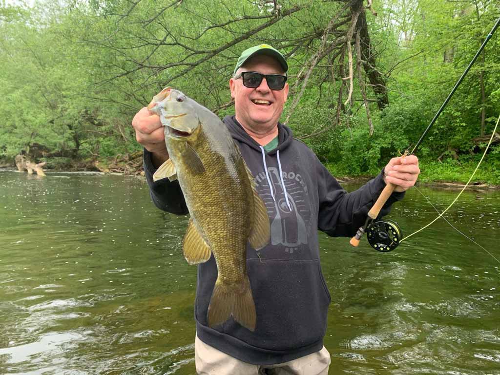 A smiling angler in sunglasses and a hat posing for a photo knee-deep in a river as he holds a Smallmouth Bass in one hand and his fly fishing rod in the other.