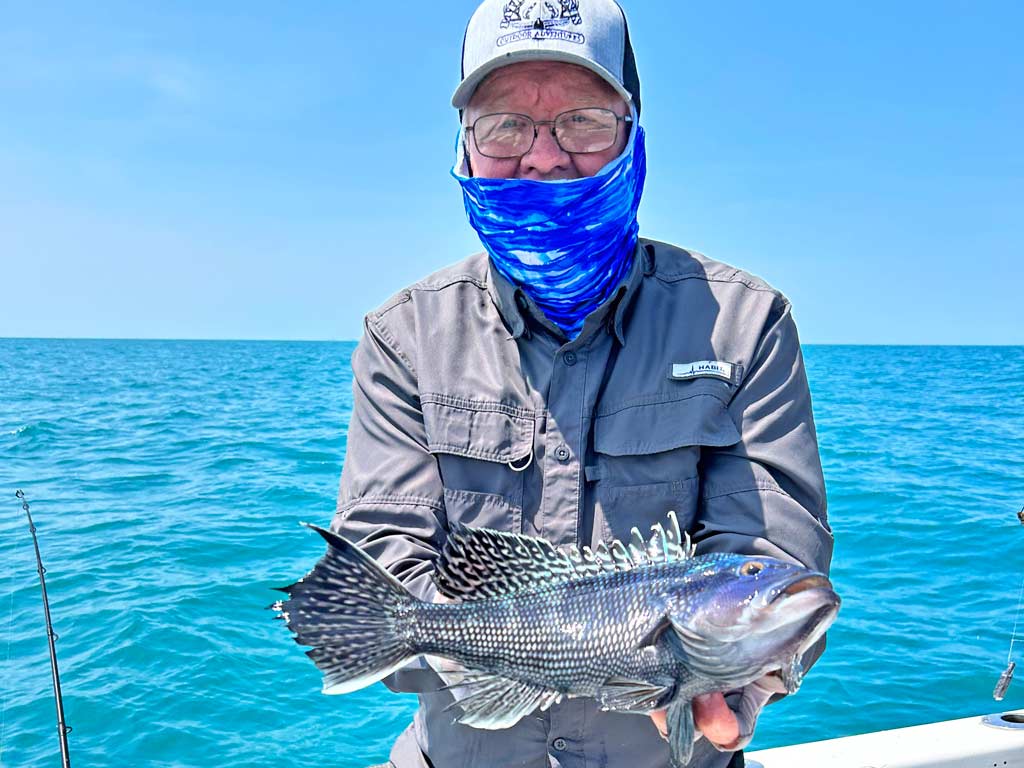 An elderly angler wearing a hat, glasses, and a bluff on his face posing for a photo on a charter boat in Ocean City, MD with a Black Seabass he caught, with electric blue waters and clear skies in the background.