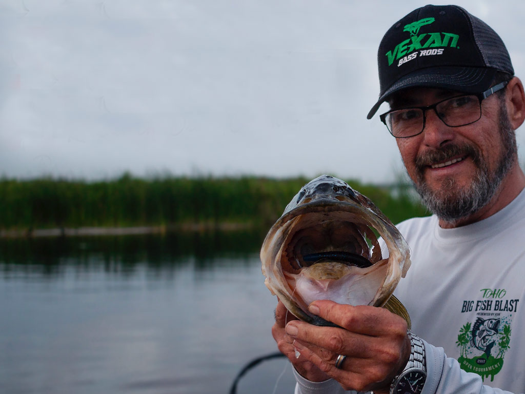 An angler wearing a hat and glasses holding a Largemouth Bass with its mouth agape towards the camera, with the bait visible deep inside the fish's mouth, along with Lake Toho's waters in the background.