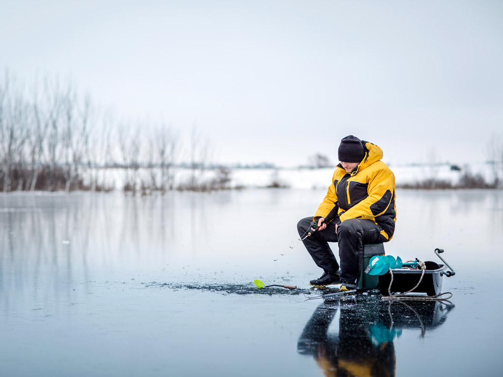 A man wearing winter clothes sitting on a bucket on a frozen lake, fishing through a hole in the ice, with a shoreline and overcast skies behind him.