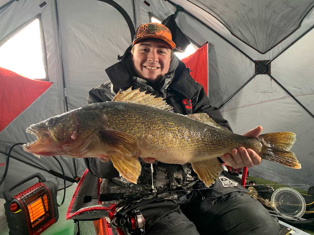 An angler sitting in an ice shanty, smiling and posing for a photo with his fishing rod across his lap and a huge Walleye in his hands, which is one of the species you can catch while ice fishing in Ohio.