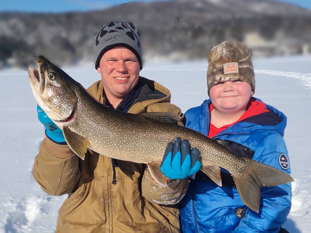 A father and a son posing for a photo on a frozen lake, with the dad holding a big Lake Trout they caught, which is one of the main targets on Schroon Lake during winter.