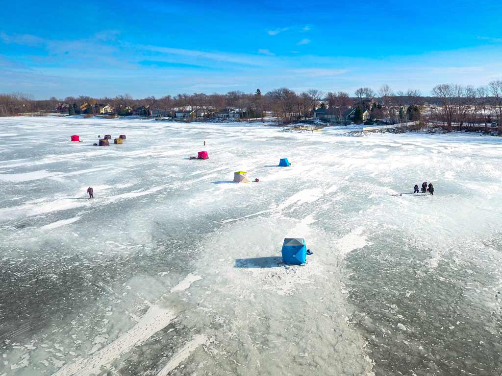 An aerial view of Green Bay in Wisconsin in winter, with several colorful ice fishing shanties propped up on the frozen waters near the shore.