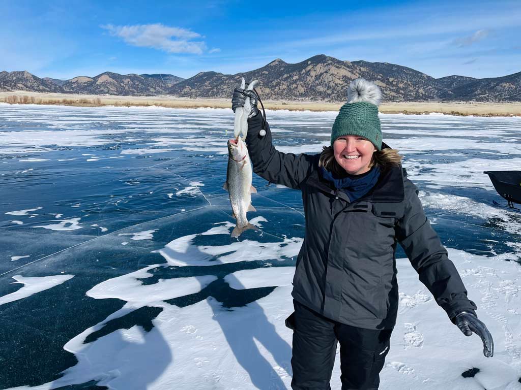 A woman wearing winter clothes and a bobble hat, standing on the frozen Eleven Mile Reservoir in Colorado and holding a Kokanee Salmon, with the ice visible behind her and mountains in the distance.